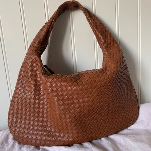 Brun axelväska i skinn - Snygg axelväska i brunt skinn med flätad design. Väskan har en mjuk, rundad form och ett stort huvudfack med dragkedja samt flera innerfack. Använd fåtal gånger💕