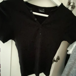 Svart topp med knappar Gina Tricot - En svart kortärmad topp från Gina Tricot i storlek XS. Toppen har en v-ringning med tre knappar framtill som ger en enkel men snygg detalj. Perfekt att matcha med jeans eller kjol för en clean look.