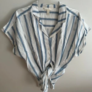 Vit och blå randig skjorta H&M - Kortärmad skjorta från H&M i vitt med blåa ränder. Skjortan har klassisk krage, knappar framtill och kan knytas nedtill för en avslappnad look. Perfekt till sommaren och passar till både jeans och shorts