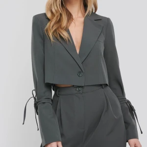 Grå croppad kavaj från NA-KD - Snygg grå croppad kavaj från NA-KD med långa ärmar och coola knyt-detaljer vid handlederna. Klassisk krage, en knapp framtill och en stilren look som passar perfekt till kostymbyxor eller jeans. 