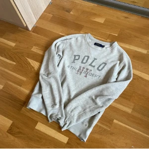 Grå sweatshirt från Polo Ralph Lauren - Grå sweatshirt från Polo Ralph Lauren med tryck på bröstet där det står 'POLO NY ATHL. DEPT.'. Klassisk rund halsringning och långa ärmar. Perfekt för en avslappnad och sportig stil.