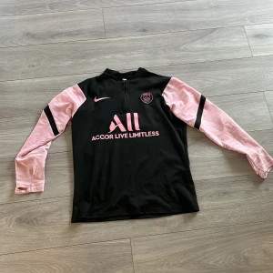 PSG långärmad träningströja Nike - Svart och rosa långärmad fotbollströja från PSG med halv dragkedja. Tröjan har rosa ärmar med svarta detaljer, PSG-logga och Nike Swoosh på bröstet samt tryck med 'Accor Live Limitless' och 'Visit Rwanda'. Tillverkad i funktionsmaterial som andas.