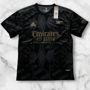 Arsenal svart matchtröja Adidas M - Snygg svart Arsenal fotbollströja från Adidas med guldiga detaljer och tryck. Tröjan har korta ärmar, rund hals och ett geometriskt mönster i svart. Tillverkad i lätt och ventilerande material med Aeroready-teknologi. Perfekt för dig som vill sticka ut på planen.