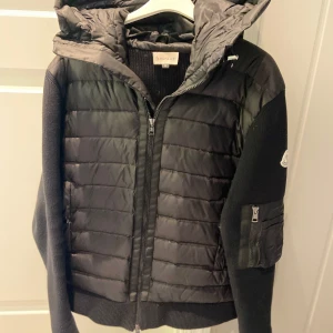 Moncler Cardigan - Snygg svart cardigan från Moncler med stickade ärmar och vadderad kropp. Jackan har huva, dragkedja framtill och en extra ficka med dragkedja på ärmen. Perfekt för dig som vill ha en stilren och exklusiv look under kyliga dagar.