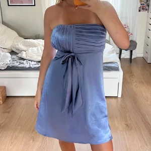  klänning med rosett - Supersöt ljusblå kort klänning i glansigt material med plisserad överdel och stor rosett i midjan. Kan bäras både axelbandslös och med smala axelband. Perfekt för sommarkvällar och studentfirande! Storlek S/M skulle jag säga (jag brukar ha storlek s)