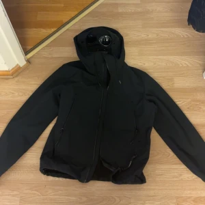 Svart Goggle Jacket från C.P. Company - Säljer en svart Goggle Jacket från C.P. Company med ikoniska glasögon i huvan. Jackan har dragkedja framtill, två snedställda fickor med dragkedja och justerbar huva. Materialet är tekniskt och känns sportigt, perfekt för dig som gillar streetwear.(pris kan diskuteras vid snabb affär) ögonen e justerbara 