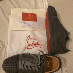 Christian Louboutin grå mocka sneakers - Snygga grå sneakers från Christian Louboutin i mocka med ikonisk röd sula. Skorna har rund tå, snörning och coola nitar framtill för en edgy vibe. Perfekt för dig som vill sticka ut med exklusiva detaljer och lyxig känsla.