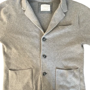 Grå cardigan jacka från Zara - Grå cardigan jacka från Zara med klassisk krage, tre knappar framtill och två stora fickor. Jackan är stickad och har en mjuk känsla, perfekt för lager-på-lager. Enkel och stilren design som funkar till många outfits.