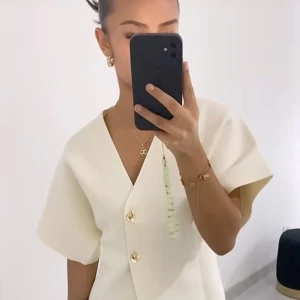 Krämvit blus med guldknappar från Zara - Minimalistisk krämvit blus från Zara med v-ringning, korta vida ärmar och två snygga guldknappar framtill. Toppens material är strukturerad polyester som ger en cool och modern känsla. Perfekt att styla med jeans eller kjol för en clean look. Blusen är i nyskick, prislappen sitter kvar