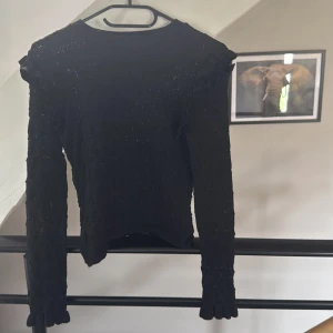 Svart stickad tröja med volang Zara - Supersnygg svart stickad tröja från Zara med långa ärmar och volangdetaljer på axlarna. Tröjan har ett diskret hålmönster som ger en cool look och passar perfekt till jeans eller kjol. Enkel rund halsringning och mjukt material.