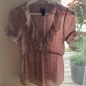 Rosa volangblus från H&M - Söt ljusrosa blus från H&M med volangkant längs v-ringningen och ärmslut. Blusen har korta puffärmar och ett subtilt mönster i tyget. Perfekt för dig som gillar romantiska detaljer och en luftig känsla.