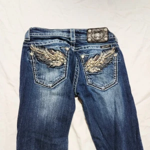 Miss Me bootcut jeans med broderi, stl 27 - Snygga blå bootcut jeans från Miss Me med broderade detaljer i guld och silver på bakfickorna. Jeansen har slitningar och kontrastsömmar, samt en cool patch med logga bak i midjan. Perfekta för dig som vill sticka ut med detaljer!