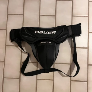 Bauer Elite Goal Jock bälte - Säljer ett Bauer Elite Goal Jock pungskydd i svart och grått. Pumgskyddet har breda, justerbara remmar och extra vaddering framtill för skydd, perfekt för hockeymålvakter.