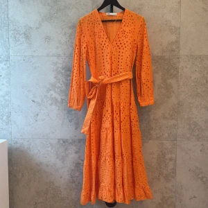 Orange långklänning från Zara - Superfin orange långklänning i bomull från Zara med broderade hålmönster, v-ringning och knytbälte i midjan. Klänningen har långa ärmar och volangdetaljer nertill, perfekt för sommarens alla tillfällen. Tvättad en gång men aldrig använd. 