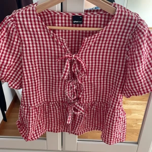 Rutig blus med knyt från Gina Tricot - Supersöt röd- och vitrutig blus från Gina Tricot med volangkant nertill, puffiga korta ärmar och tre knyt framtill. Blusen har en luftig passform och är gjord i bomull, perfekt för en somrig look. Totalt oanvänd💗