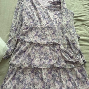 Ljuslila blommig långklänning från Vila - Superfin långklänning från Vila i ljuslila och vitt med blommigt mönster. Klänningen har långa ärmar, v-ringning och volangdetaljer. Perfekt för dig som gillar romantisk och feminin stil. Materialet är lätt och flowy, vilket gör den extra bekväm.