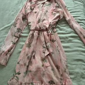 Blommig rosa kortklänning Trendyol - Supersöt rosa kortklänning från Trendyol med blommigt mönster, v-ringning och långa ärmar. Klänningen har omlottliv, volangdetalj nertill och knyt i midjan. Perfekt för vår och sommar när du vill känna dig extra fin.