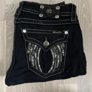 Svarta Miss Me jeans - Svarta jeans från Miss Me med kontrasterande vita sömmar och snygga detaljer. Bakfickorna har broderade vingar och glittrande stenar, samt dekorativa knappar med mönster. Klassisk femficksmodell och logga på linningen.
