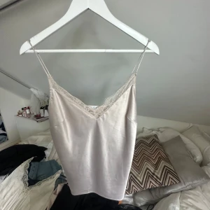 Champagnefärgad linnetopp med spets - Snygg champagnefärgad linnetopp med tunna axelband och v-ringning dekorerad med spets. Toppen har en silkeslen känsla och en enkel, elegant look som passar perfekt till jeans eller kjol.