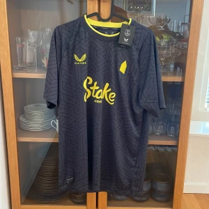 Everton bortatröja 24-25 från Castore - Snygg Everton FC bortatröja 2024-2025 från Castore i mörkgrått med geometriskt mönster och gula detaljer. Tröjan har korta ärmar, rund hals med gul kant och tryckta loggor. Tillverkad i lätt och ventilerande funktionsmaterial. Samlar själv på fotbollströjor och kan garantera att Vara är 100% äkta