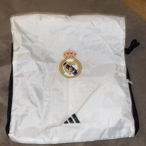 Vit Real Madrid gympåse - Säljer en vit gympåse med Real Madrid-logga och Adidas-märke. Påsen har snörning i svart och en dragkedja på sidan för extra förvaring. Köpt i spanien i real madrids affär. Knappt använd