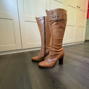 Bruna höga boots från Roots, storlek 39 - Snygga höga boots från Roots i brunt skinn med bred klack och dekorativt spänne upptill. Dragkedja på insidan för enkel påtagning. Perfekta för dig som vill ha en klassisk men trendig look.