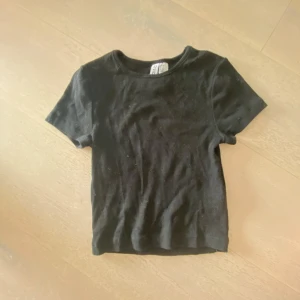 Svart ribbad topp från Divided XS - En svart, ribbad topp från Divided i storlek XS. Modellen är croppad med korta ärmar och rund halsringning. Perfekt för dig som gillar en enkel och stilren look. Materialet är mjukt och stretchigt för en bekväm passform.