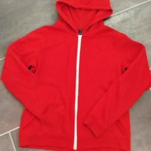 Röd hoodie med vit dragkedja - Riktigt snygg röd hoodie med huva och vit dragkedja framtill. Tröjan har långa ärmar och är tillverkad i mjukt material, perfekt för att chilla eller dra på sig när det är lite kyligt. Klassisk passform och enkel stil som funkar till det mesta.