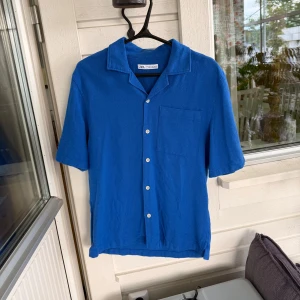 Blå kortärmad skjorta från Zara - Snygg blå skjorta från Zara i storlek M. Skjortan har korta ärmar, klassisk krage, bröstficka och vita knappar framtill. Tillverkad i mjuk bomulls- och polyestermix som känns skön mot huden. Perfekt för en chill och stilren look.