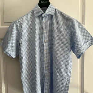 Blåvit rutig kortärmad skjorta - Snygg blåvit rutig skjorta från Herr Karl med korta ärmar och klassisk krage. Skjortan har knappar framtill och är tillverkad i mjuk bomull, perfekt för varma dagar. Passar dig som gillar stilren och fräsch look.