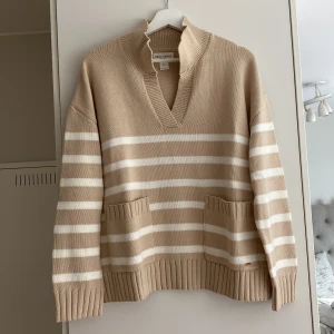 Beige och vit randig stickad tröja - Superfin stickad tröja från Holly & Whyte i beige och vitt med breda ränder. Tröjan har v-ringad krage, två stora fickor framtill och ribbade muddar. Perfekt oversized passform för en chill och trendig look.