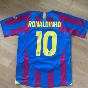 Barcelona Ronaldinho 10 CL Final 2006 tröja - Säljer en ikonisk FC Barcelona fotbollströja från Champions League-finalen 2006 mot Arsenal. Tröjan är randig i blått och rött med gula detaljer, Ronaldinho och nummer 10 på ryggen, samt broderad finaltext på ärmen. Klassisk Nike-logga och klubbmärke på bröstet. Perfekt för samlare!