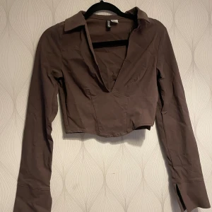 Brun croppad långärmad topp från H&M Divided - Säljer en brun croppad långärmad topp från H&M Divided. Toppen har en djup v-ringning och krage, vilket ger en trendig look. Perfekt att matcha med jeans eller kjol för en snygg outfit.