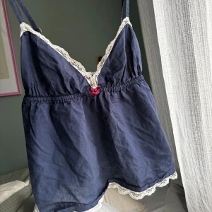 Odd Molly nattlinne - Söt mörkblå linnetopp/nattlinne med smala axelband och dekorativ vit spets längs urringning och benslut. Framtill sitter en liten röd rosett. Toppen har en avslappnad passform och resår i midjan. Storlek 0
