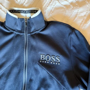 Mörkblå zip-jacka från Hugo Boss - Säljer en mörkblå zip-hoodie från Hugo Boss med   har vita detaljer vid dragkedjan och på kragen.  Nyskick nästintill oanvänd. Storlek : S
