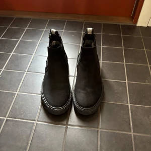 Svarta chelsea boots från Vagabond - Säljer ett par svarta chelsea boots från Vagabond med elastiska paneler på sidorna och dragflik bak. Skorna har rund tå och platt sula, tillverkade i mocka för en stilren look.
