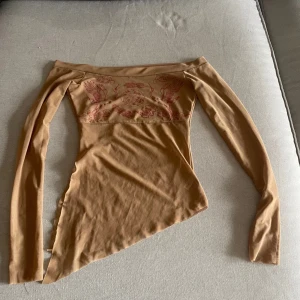Vintage Beige offshoulder topp med rosa tryck - Säljer en beige offshoulder topp med långa ärmar och rosa tryck av tranor och moln över bröstet. Toppen har asymmetrisk nederkant och är figurnära i modellen. Perfekt för dig som gillar unika detaljer och stilren design.