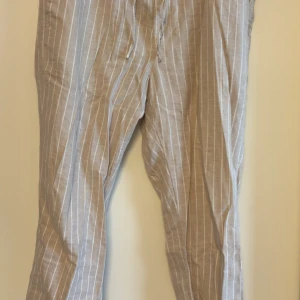 linnebyxor  - Säljer ett par beige linnebyxor från H&M med vita vertikala ränder. Byxorna har normal passform och dragsko i midjan för extra komfort. Perfekta för varmare dagar och en avslappnad stil.