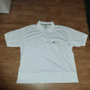 Vit piké t-shirt från Lacoste - Klassisk vit piké t-shirt från Lacoste med krage, knappar och bröstficka. Ikoniska krokodilloggan på bröstet och ärmen. Perfekt för en stilren och sportig look.