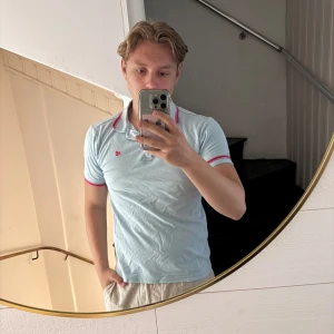 Ljusblå piké från Giogoi - Säljer en ljusblå pikétröja från GoGo med rosa detaljer på krage och ärmslut. Tröjan har korta ärmar och en liten broderad logga på bröstet. Perfekt för en avslappnad och stilren look.