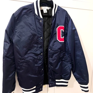 Blå bomberjacka  - Snygg blå bomberjacka från Zara med vit och svart ribbad krage, ärmslut och nederkant. Stor röd och vit C-patch på bröstet ger en sportig look. Jackan har knappar framtill och quiltat foder. 