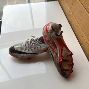 Nike Mercurial fotbollsskor i vitt och rött - Säljer ett par Nike Mercurial fotbollsskor i vitt med svarta detaljer och röd sula. Skorna säljs eftersom de bara har legat runt efter att de har blivit för små. Skorna är i bra skick men bara lite smutsiga