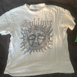 Vit t-shirt med Sublime-tryck - Vit oversize t-shirt med stort tryck av Sublime-loggan och ett solansikte på framsidan. Klassisk passform med rund hals och korta ärmar. Har några små hål framtill och även på sidan från etiketten vid köp annars fräsch!