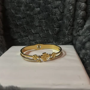 Guldfärgat armband med fyrklöver och stenar - Snyggt guldfärgat armband med dekorativa fyrklöver och små gnistrande stenar. Armbandet har en elegant och stilren design som passar perfekt till att lyfta din outfit. Rostfritt!!