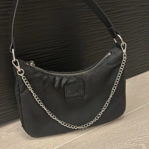 Svart axelväska med kedjedetalj - Snygg svart axelväska med silverfärgad kedja och dragkedja upptill. Väskan har en stilren design med ett litet märke framtill och ett rymligt huvudfack. Perfekt för att bära dina viktigaste saker.