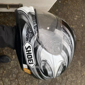 svart&vit motorcykelhjälm/mopedhjälm från Shoei - Säljer en snygg motorcykelhjälm/mopedhjälm från Shoei i svart och vitt med gråa detaljer och grafiskt mönster. Hjälmen har visir och klassisk rund form. Perfekt för dig som vill ha både stil och säkerhet på vägen. Kan gå ner i pris