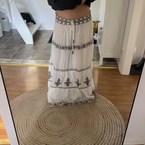 Vit långkjol med broderade detaljer och knytband - Säljer en vit långkjol med mörkgrå/mörkbeige /svart broderade mönster och dekorativa band. Kjolen har ett knytband i midjan med pärlor och är luftig, jag är 160 så kjolen är lite lång på mig den är storlek L men passar även M och S