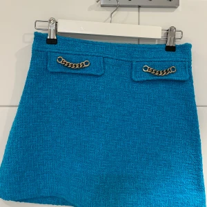 Turkos kjol med kedjedetaljer från ZARA - Säljer en turkos kortkjol från ZARA i strukturerat tyg. Kjolen har två dekorativa fickor framtill med guldfärgade kedjor som detalj. Perfekt för dig som vill sticka ut med färg och snygga detaljer.