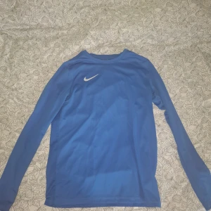 Blå långärmad träningströja från Nike - Säljer en blå långärmad träningströja från Nike med vit logga på bröstet. Tröjan är i slim fit-modell och tillverkad i Dri-FIT-material som andas, perfekt för sport och träning.