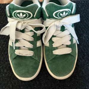 Adidas Campus gröna sneakers - Säljer ett par gröna Adidas Campus sneakers med vita ränder och vit sula. Skorna har breda vita skosnören och är tillverkade i mocka med klassisk Adidas-logga på plös och häl. Perfekta för dig som gillar retrostil och streetwear. Priset kan diskuteras 